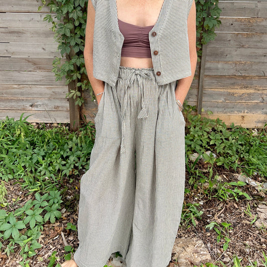 A person standing in front of a wooden fence and greenery, wearing wide legged, high waisted trousers with a drawstring waist in a black and white pin stripe pattern.