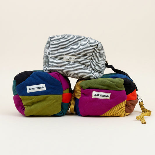 Three small fabric bags from Dear Friend stacked. The top bag is striped black and white, while the two bags below are made of colorful patchwork fabric in purple, green, blue, yellow, and red.
