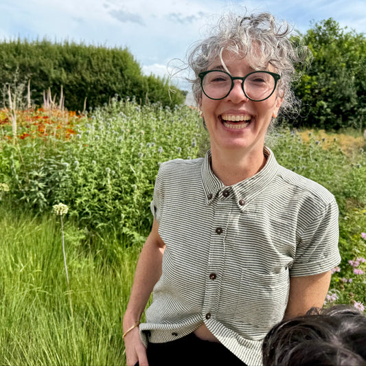 A smiling woman with short gray curly hair and black-rimmed glasses stands outdoors in a lush green garden, wearing a black and white striped short-sleeve button-up shirt.