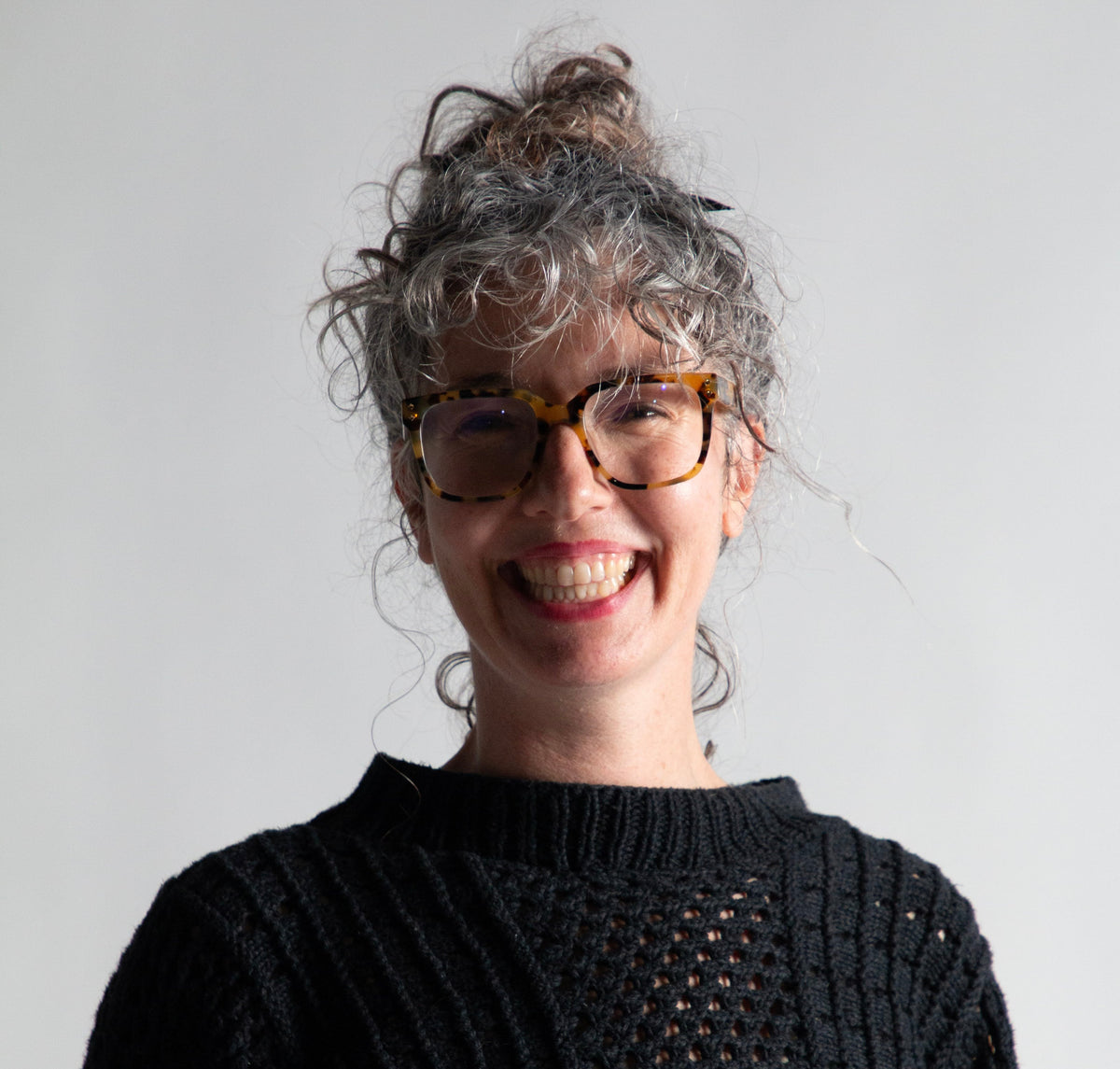 A smiling woman with curly gray hair and tortoiseshell glasses wearing a black cable-knit sweater, photographed against a plain light gray background.