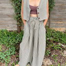 A person standing in front of a wooden fence and greenery, wearing wide legged, high waisted trousers with a drawstring waist in a black and white pin stripe pattern.