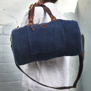 A person carrying a blue mini duffel bag with brown leather trim and a canvas shoulder strap.