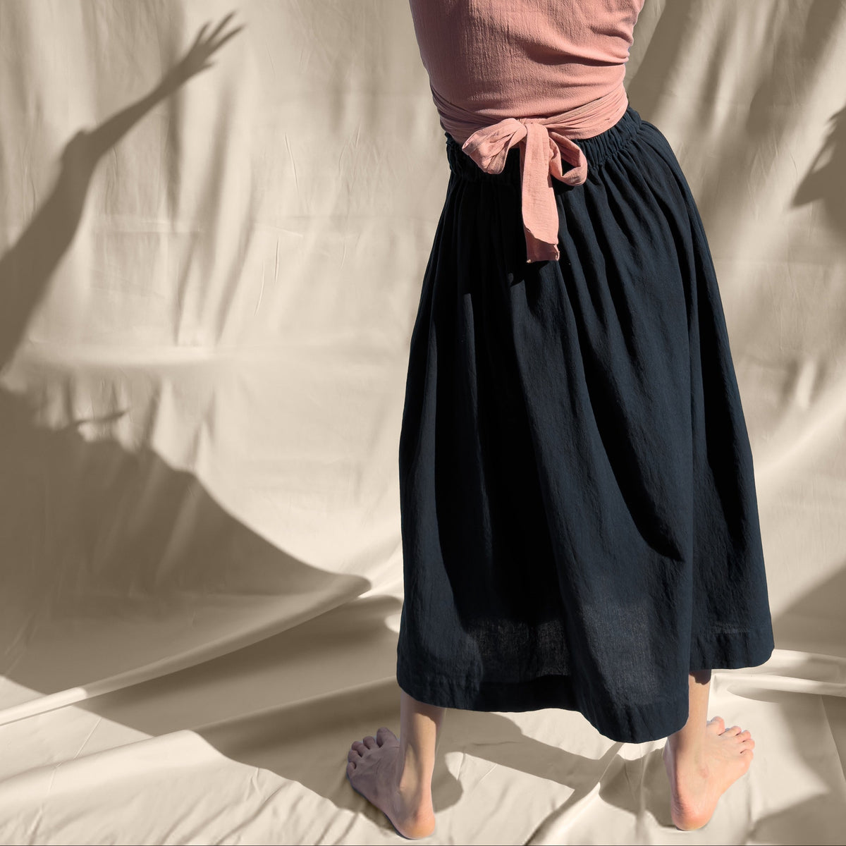 Person in a pink top and black skirt standing with arms raised against a white curtain.