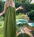 A woman wearing a long green spaghetti-strap maxi dress and a wide-brimmed straw hat stands in a garden setting with a green metal table and chairs.