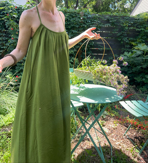 A woman wearing a long green spaghetti-strap maxi dress and a wide-brimmed straw hat stands in a garden setting with a green metal table and chairs.