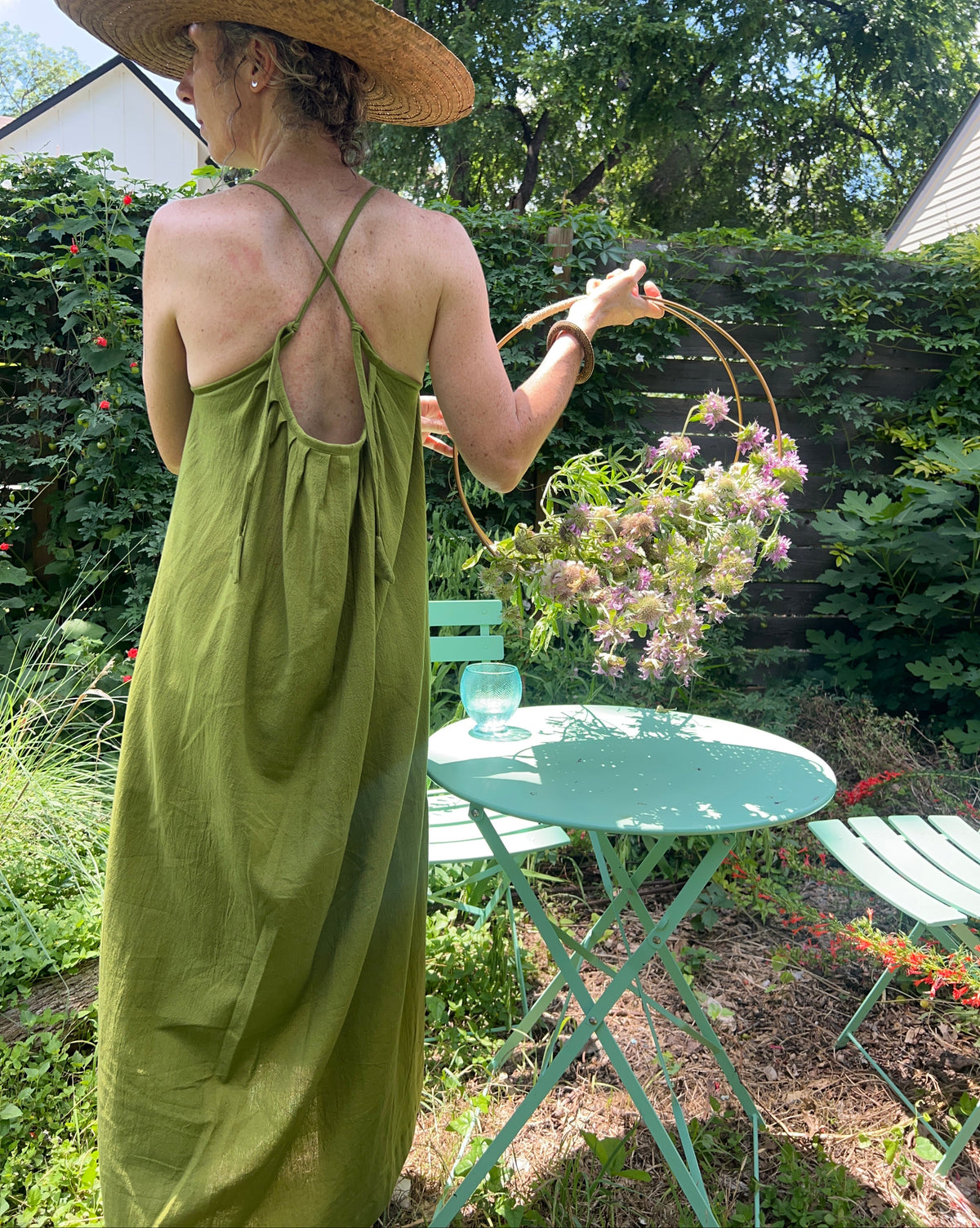 A woman wearing a wide-brimmed straw hat and a green maxi dress stands in a garden, holding a large circular wire frame. A mint green metal patio table with a glass and a flower arrangement is in the foreground.