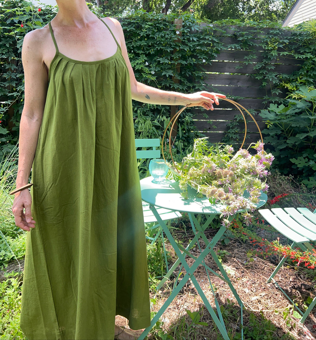 A woman wearing a wide-brim straw hat and a green sleeveless maxi dress stands in a garden next to a mint green folding table with a flower basket.