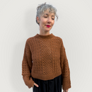A woman with short gray curly hair wearing a textured brown knit sweater and dark pants, standing against a plain light gray background.