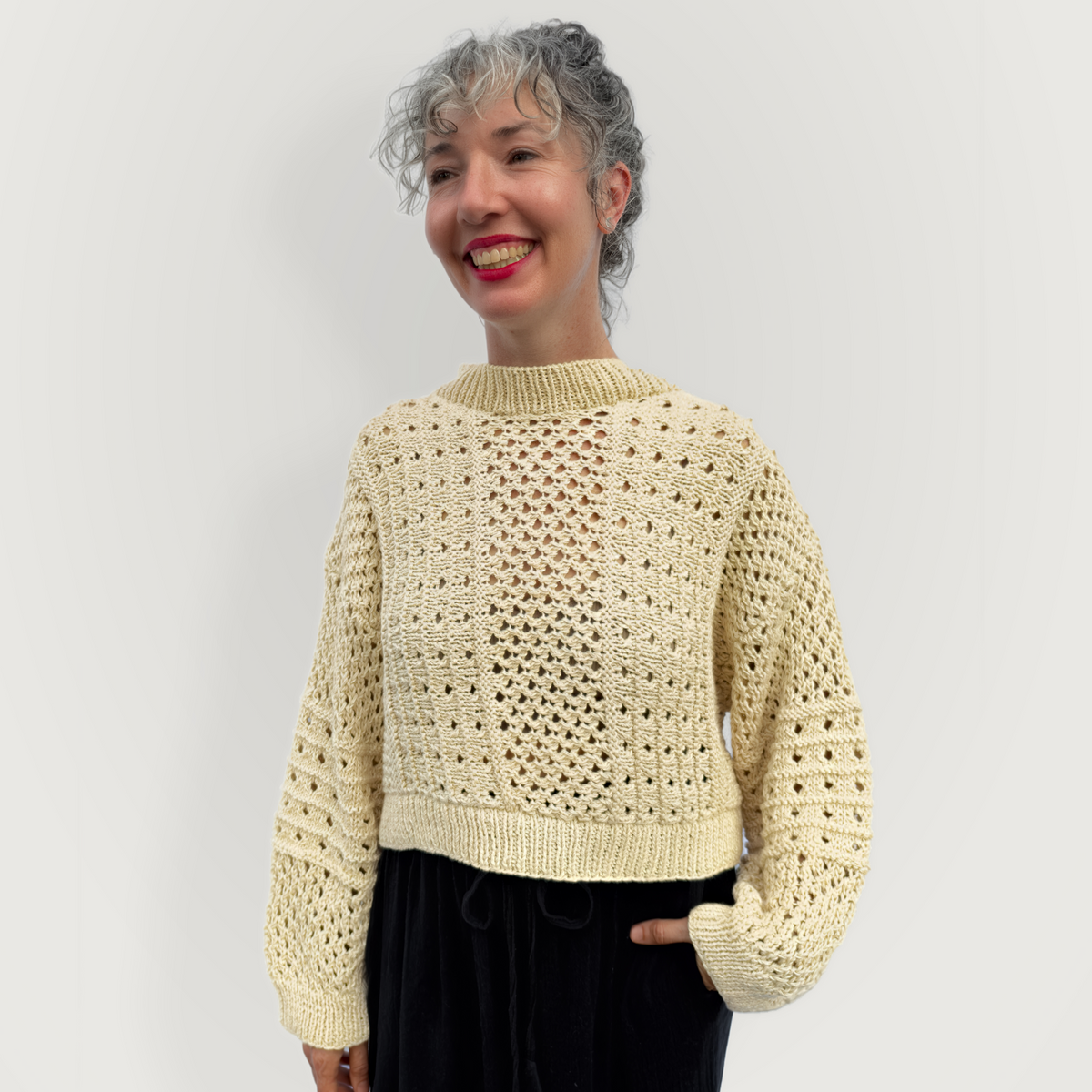 A woman with gray hair smiling and wearing a cream-colored, open-knit cropped sweater with long sleeves, paired with black pants, against a plain light gray background.
