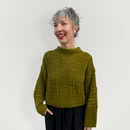 A woman with gray hair smiling and looking to the side, wearing a textured olive green knit sweater and black pants, standing against a plain light gray background.