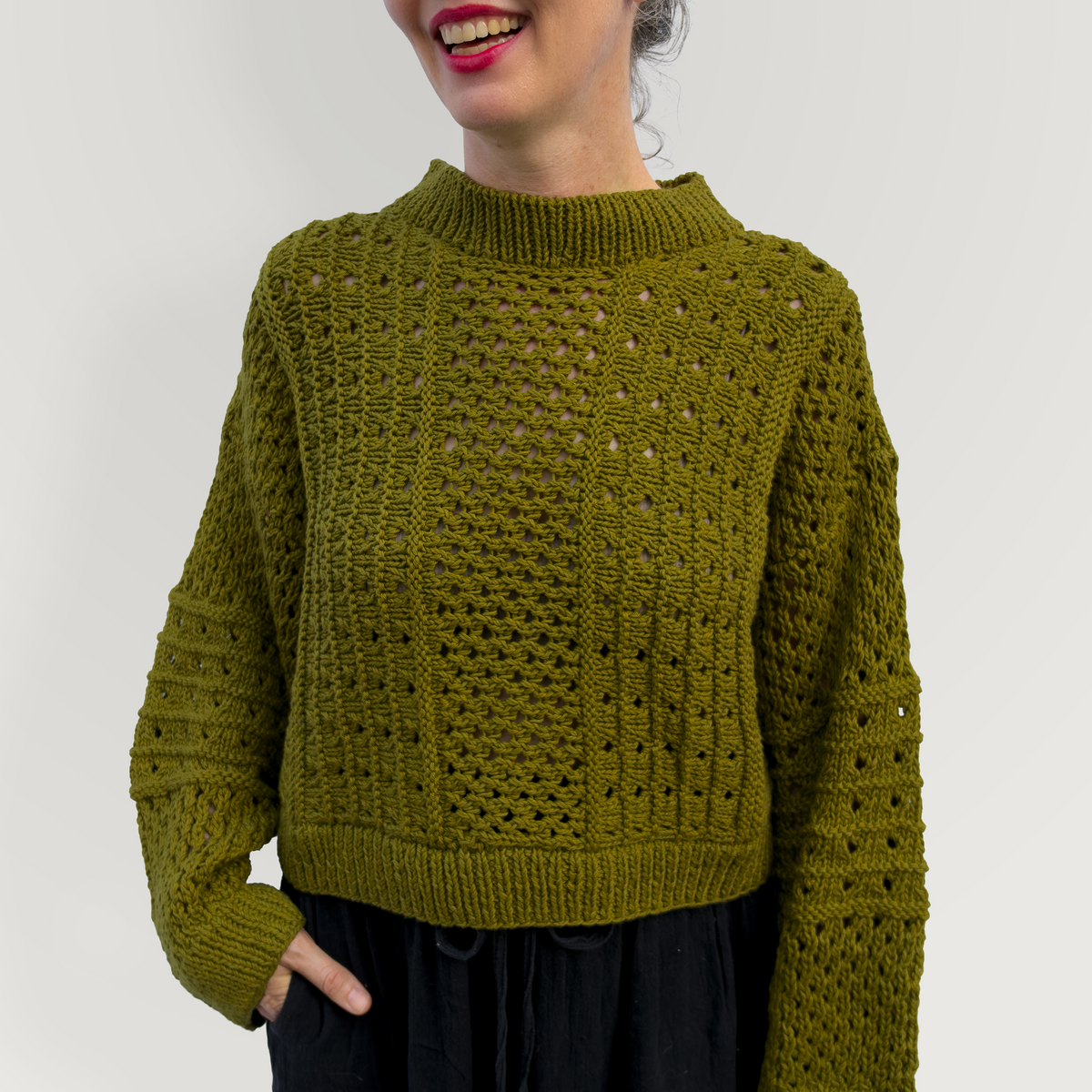 A woman with gray hair smiling and looking to the side, wearing a textured olive green knit sweater and black pants, standing against a plain light gray background.