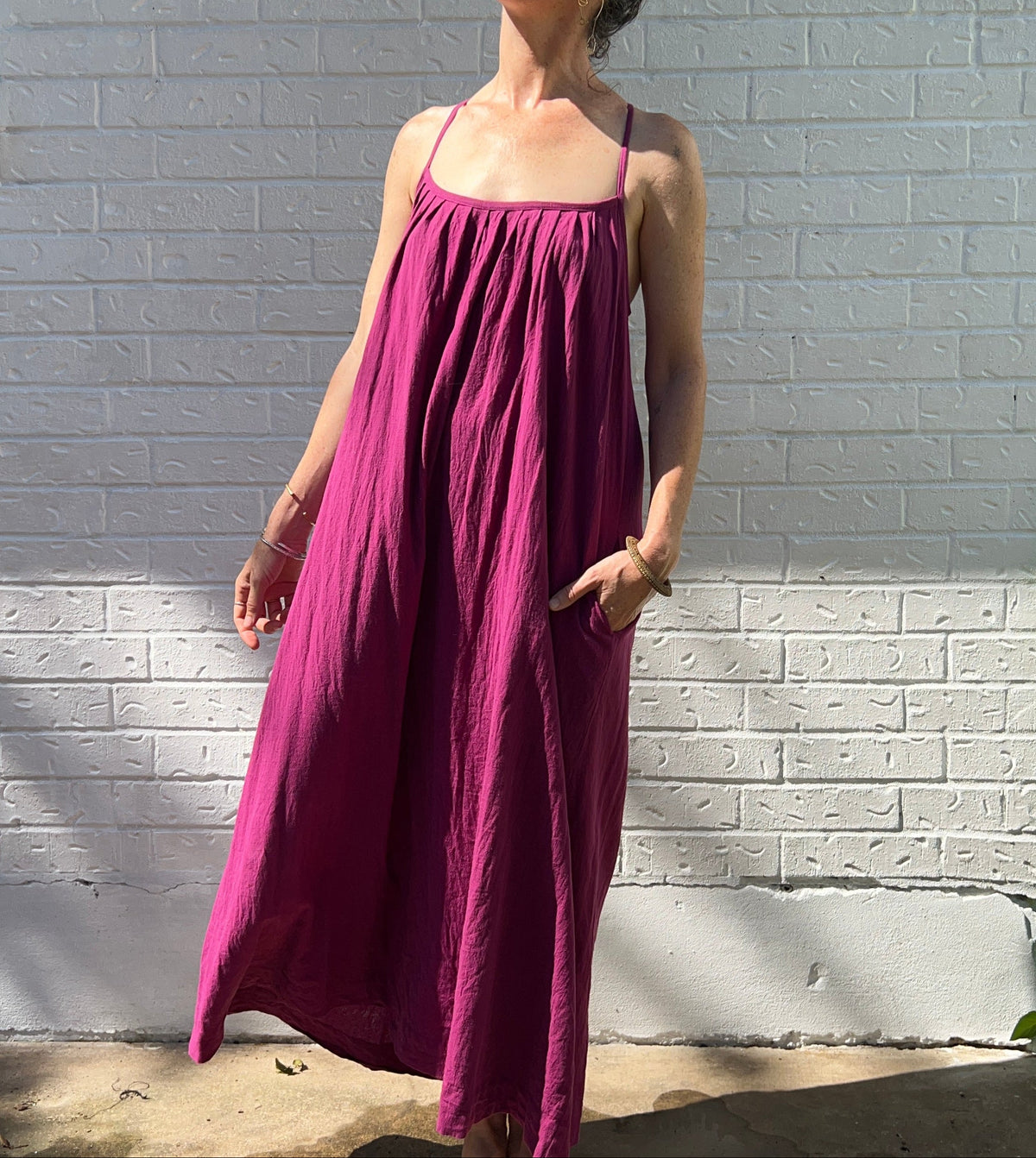 A woman wearing a sleeveless magenta maxi dress with pockets, standing barefoot against a white painted brick wall outdoors.