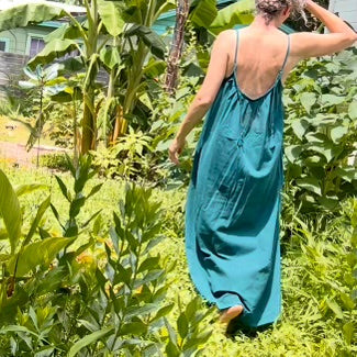 A woman wearing a teal blue maxi slip dress walks through a lush green garden, viewed from behind to show the back of the dress with its thin straps and open back.