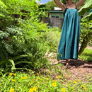 A woman wearing a teal blue-green linen slip dress stands in a lush garden with her arms raised, surrounded by green plants, yellow flowers, and a wooden fence in the background.