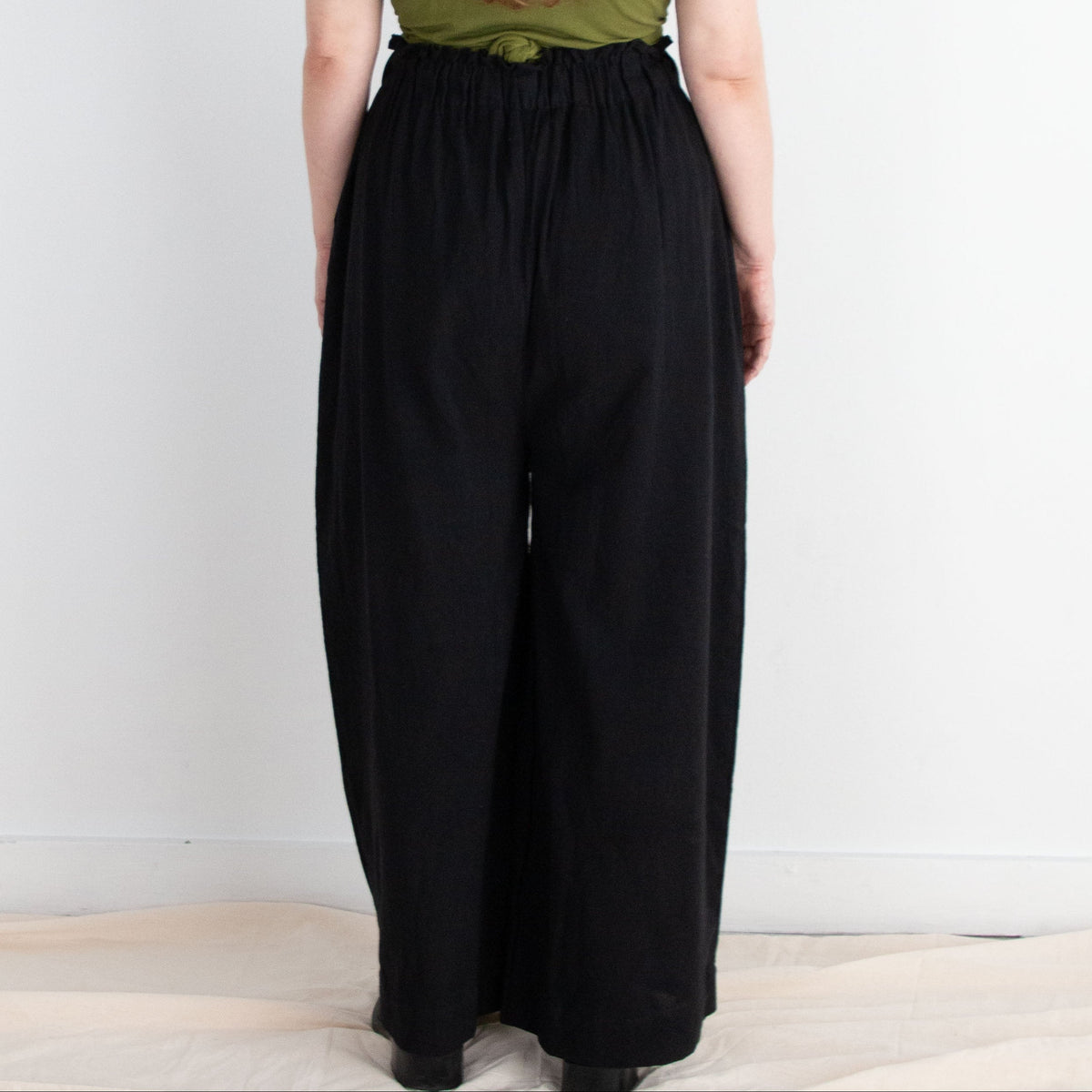 Person wearing a green top and black pants standing against a white wall.
