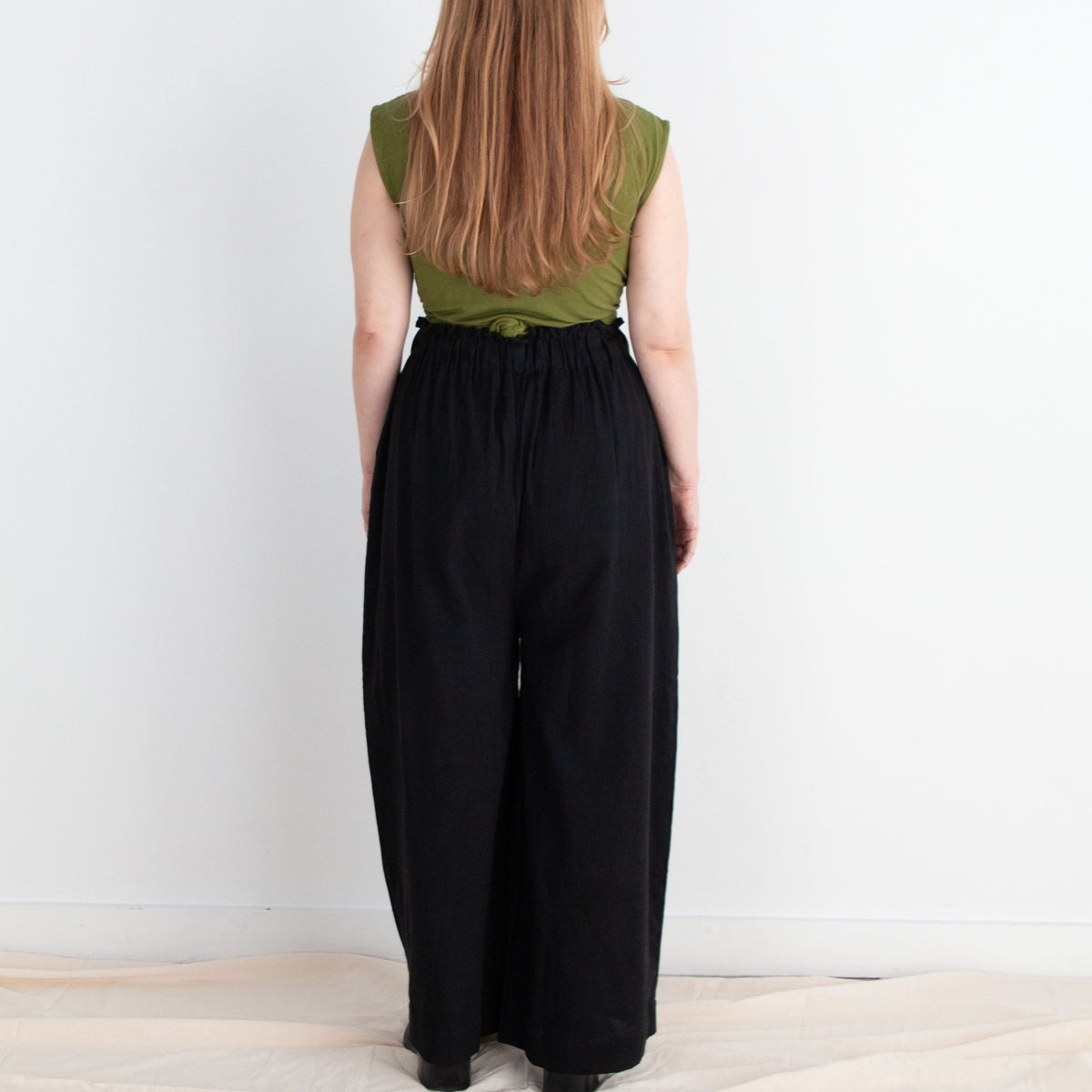 Person wearing a green top and black pants standing against a white wall.