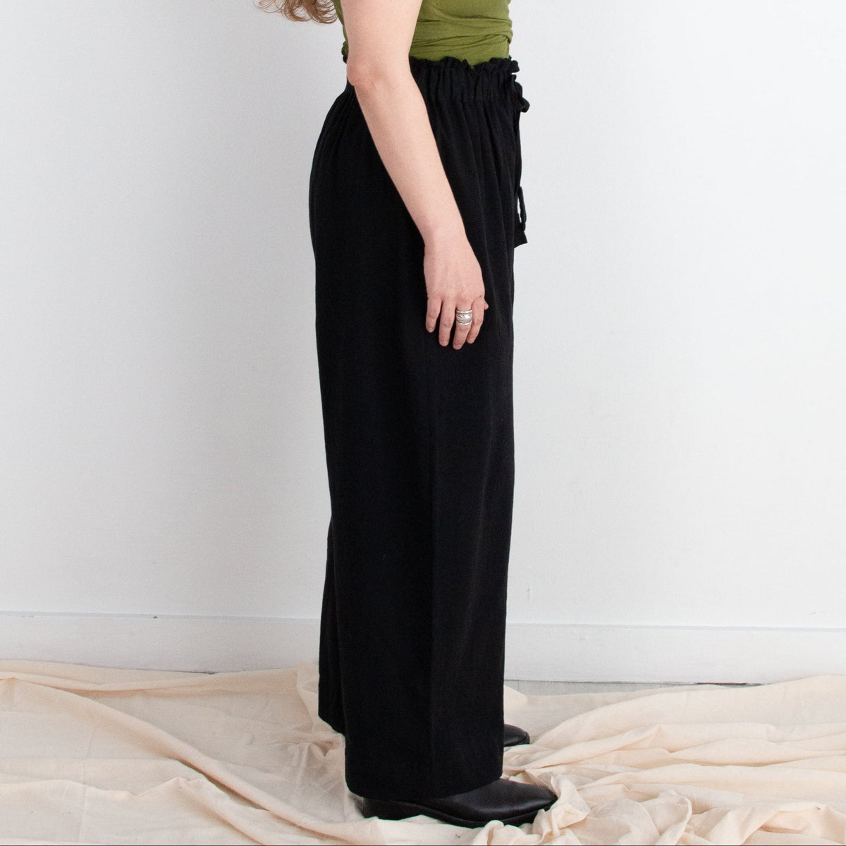 Woman wearing a green top and black pants standing against a white wall with draped fabric on the floor.