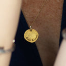 Gold necklace with a round disk, written "here & now", on a person wearing a dark top.