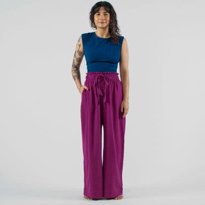 Woman wearing a blue sleeveless top and purple wide-leg pants standing on a white background.