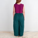 Woman wearing a purple top and teal pants standing against a white wall.