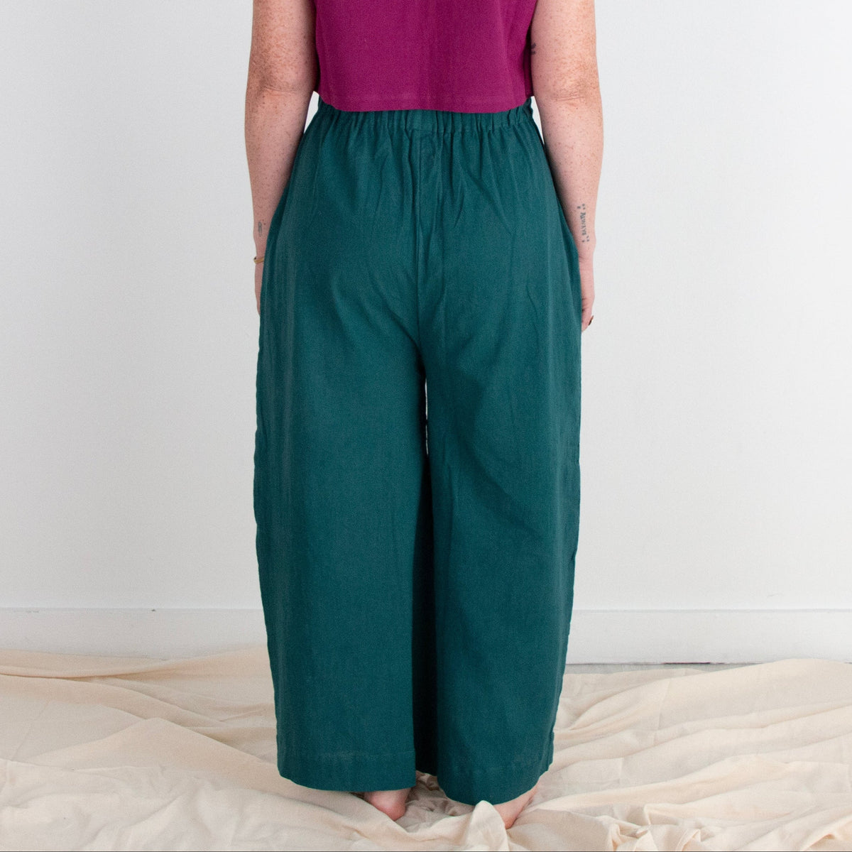 Woman wearing a purple top and teal pants standing against a white wall.