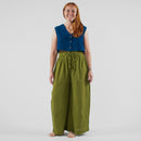 Woman wearing a blue sleeveless top and green wide-leg pants standing in front of a white backdrop with photography equipment.