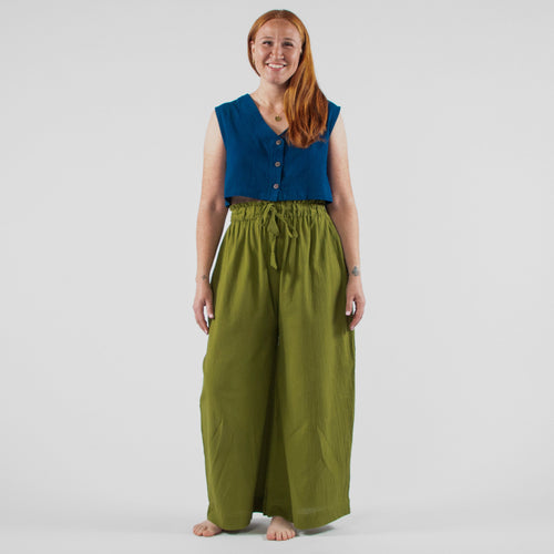 Woman wearing a blue sleeveless top and green wide-leg pants standing in front of a white backdrop with photography equipment.