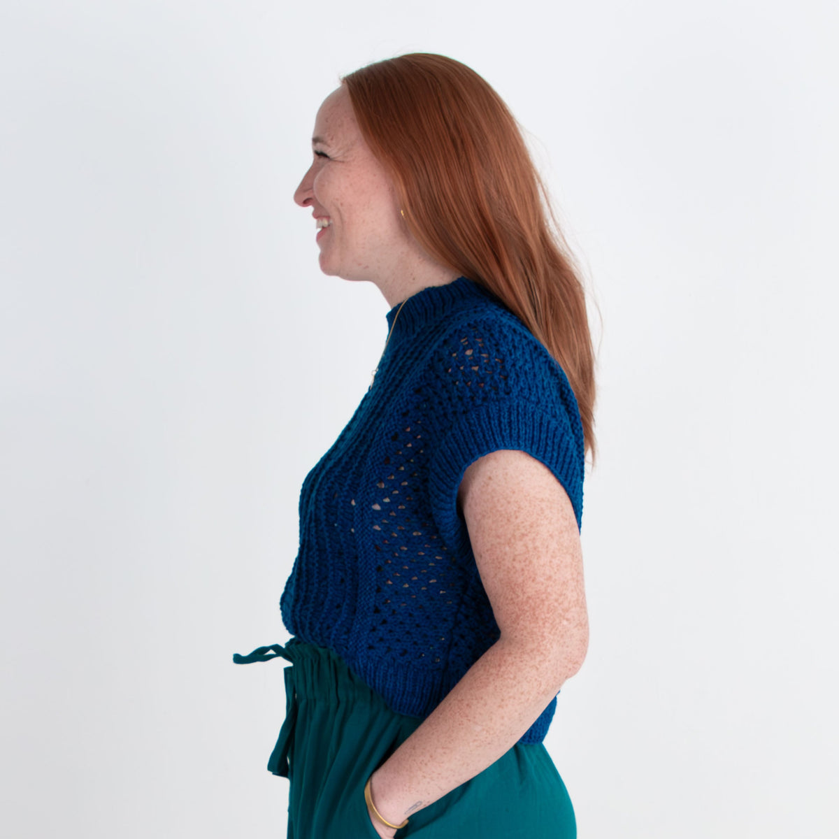 Woman wearing a blue top and teal pants standing against a white background