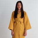 A woman wearing a mustard yellow striped wrap dress with a tie belt, standing against a plain white wall.