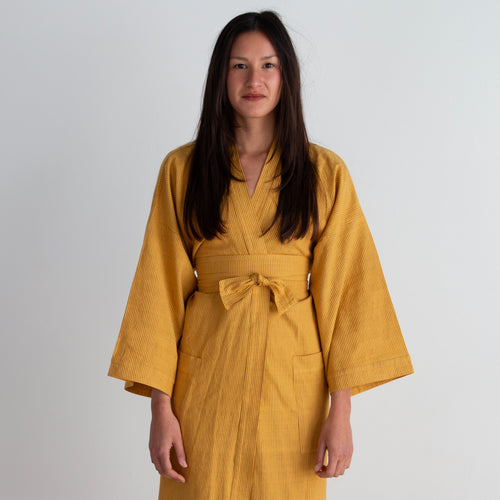 A woman wearing a mustard yellow striped wrap dress with a tie belt, standing against a plain white wall.