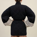 A person shown from the back wearing a black quilted puffer jacket with beige knit sleeve inserts, posed against a white fabric backdrop.