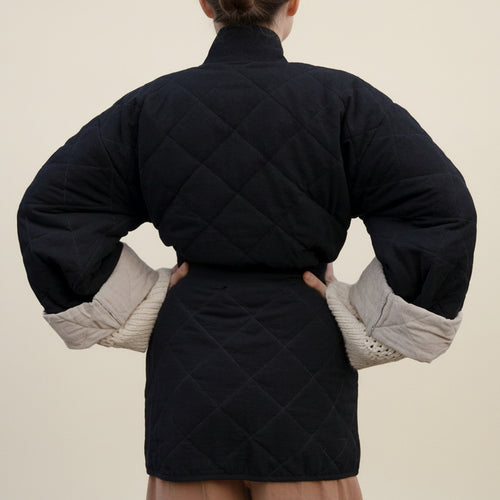 A person shown from the back wearing a black quilted puffer jacket with beige knit sleeve inserts, posed against a white fabric backdrop.