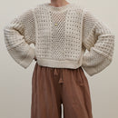 A person wearing a chunky cream open-knit cropped sweater with voluminous sleeves and brown drawstring pants, photographed against a plain light beige studio background.