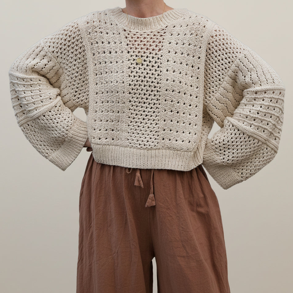 A person wearing a chunky cream open-knit cropped sweater with voluminous sleeves and brown drawstring pants, photographed against a plain light beige studio background.