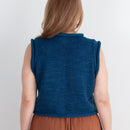 Woman wearing a blue sleeveless top and brown pants standing against a white wall.