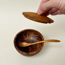 A small round wooden bowl with a matching wooden spoon inside, a hand lifts the lid above it.
